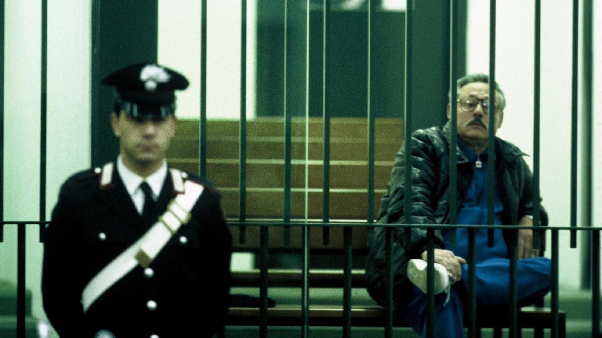 "Italiens Mafia - Paten vor Gericht": Ein Mann sitzt hinter Gittern. Davor steht ein Polizist.