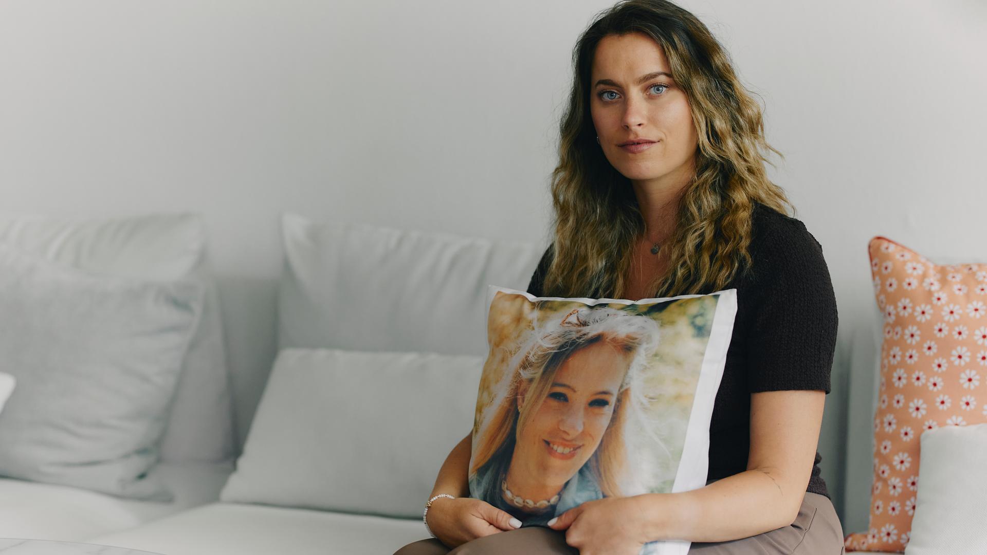 Jacky sitzt auf einer Couch und schaut straight in die Kamera. In ihren Händen hält sie ein Kissen, auf dem ein Foto ihrer vertorbenen Zwillingsschwester zu sehen ist.