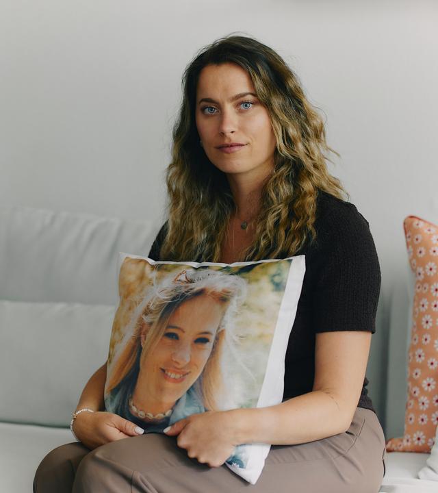 Jacky sitzt auf einer Couch und schaut straight in die Kamera. In ihren Händen hält sie ein Kissen, auf dem ein Foto ihrer vertorbenen Zwillingsschwester zu sehen ist.