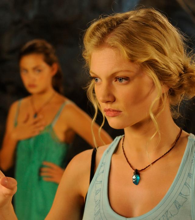 Rikki steht mit angestrengtem Blick nach vorne in einer Höhle. Ihre rechte Hand hat sie zu einer Kralle geballt. Im Hintergrund stehen Bella und Cleo, die Rikki besorgt anschauen.