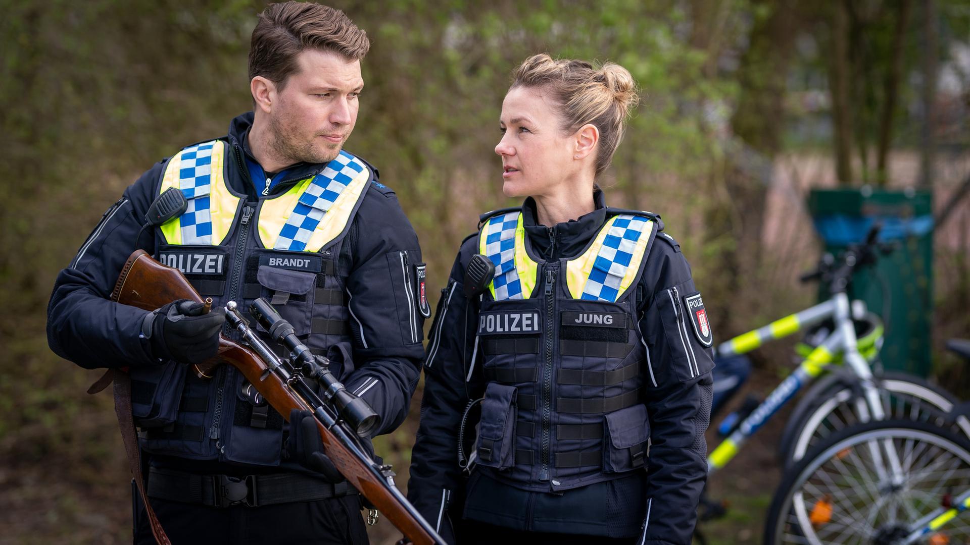 "Notruf Hafenkante - Jagdzeit": Der Polizist Nick Brandt (Raúl Richter) und seine Kollegin Franzi Jung (Rhea Harder-Vennewald) stehen dicht nebeneinander in einem Park. Nick hält ein Gewehr in den Händen. Beide sehen sich an. Im Hintergrund sind zwei abgestellte Fahrräder zu erkennen.