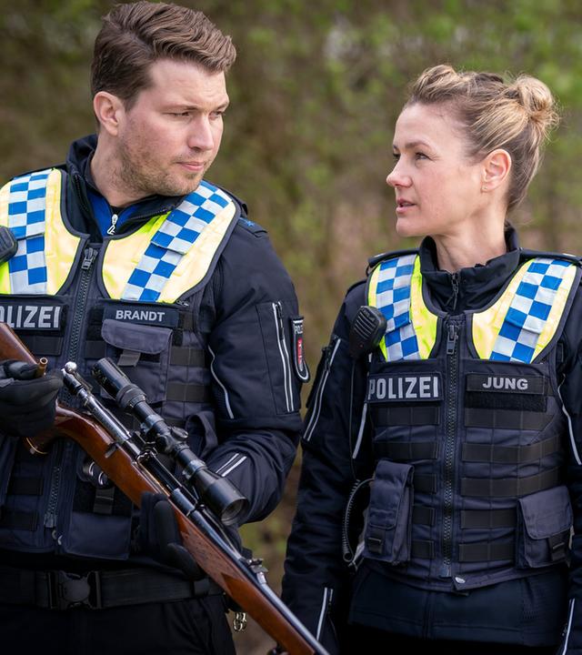 "Notruf Hafenkante - Jagdzeit": Der Polizist Nick Brandt (Raúl Richter) und seine Kollegin Franzi Jung (Rhea Harder-Vennewald) stehen dicht nebeneinander in einem Park. Nick hält ein Gewehr in den Händen. Beide sehen sich an. Im Hintergrund sind zwei abgestellte Fahrräder zu erkennen.