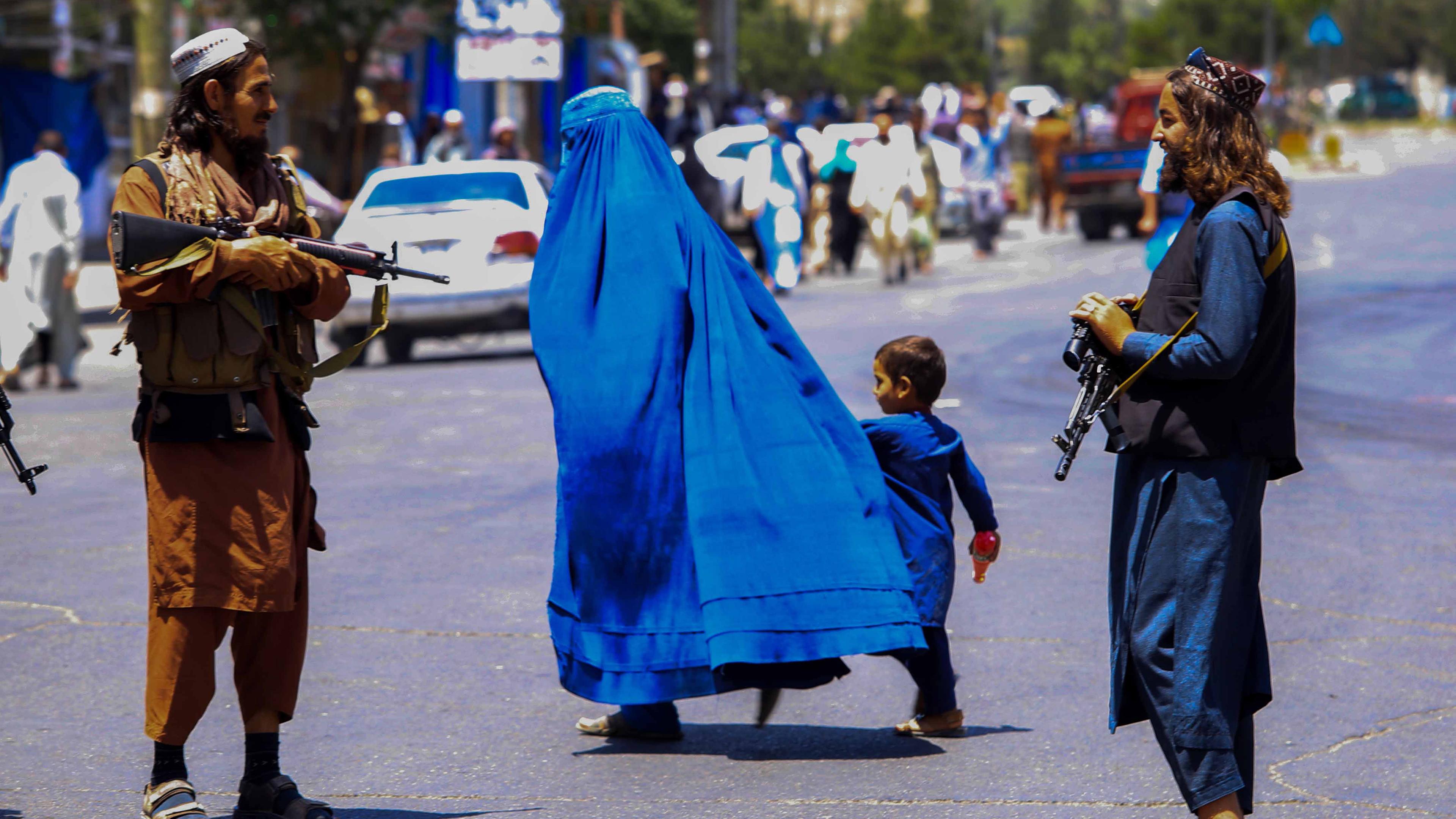 Ein Sicherheitsmann der Taliban steht bewaffnet auf offener Straße neben einer Frau mit ihrem Kind.