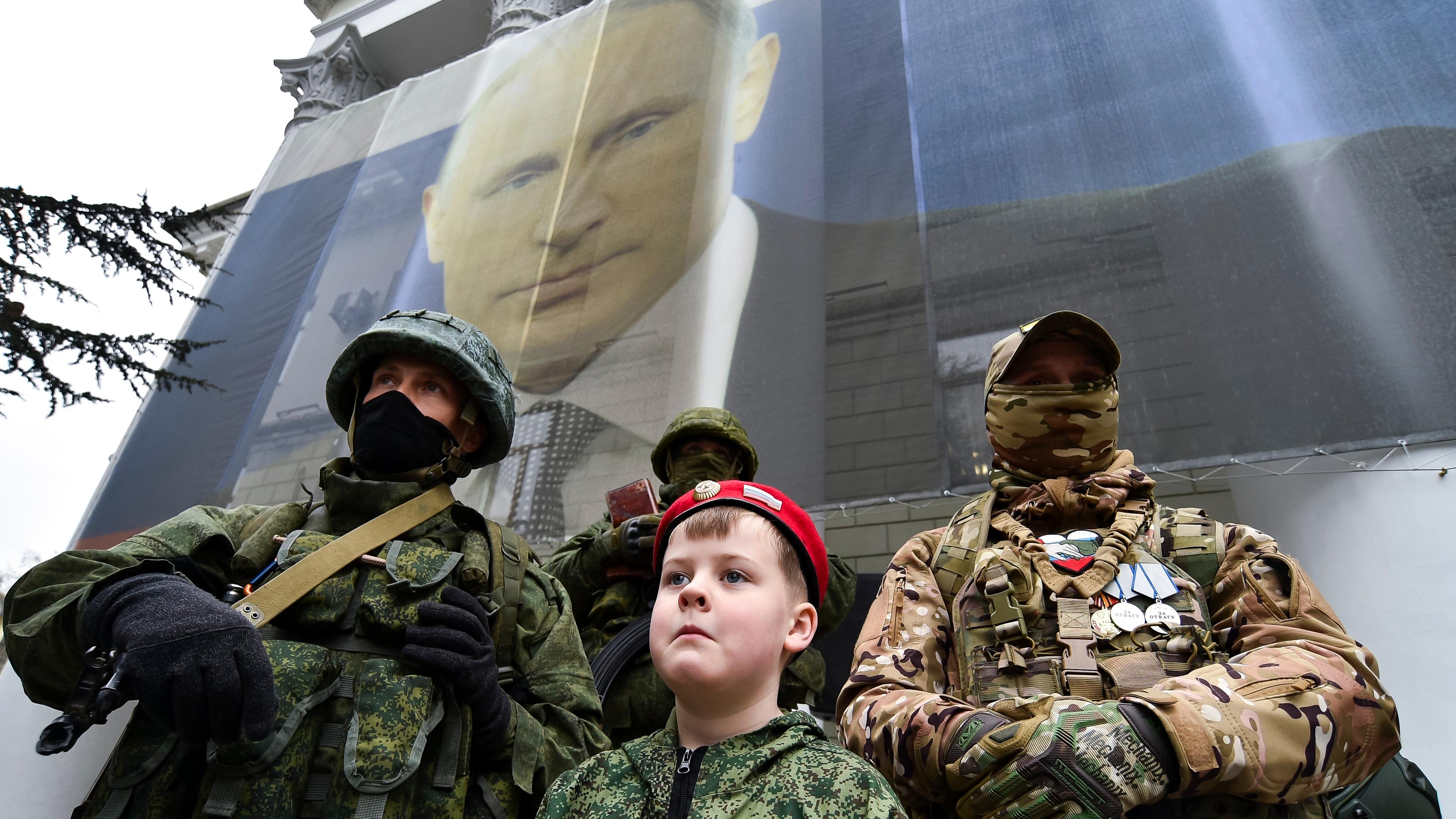 Ein Junge steht vor zwei russischen Soldaten, im Hintergrund ist das Gesicht Putins auf eine Hauswand gemalt. Die Soldaten und der Junge nehmen an einer Aktion zum neunten Jahrestag der Krim-Annexion teil.
