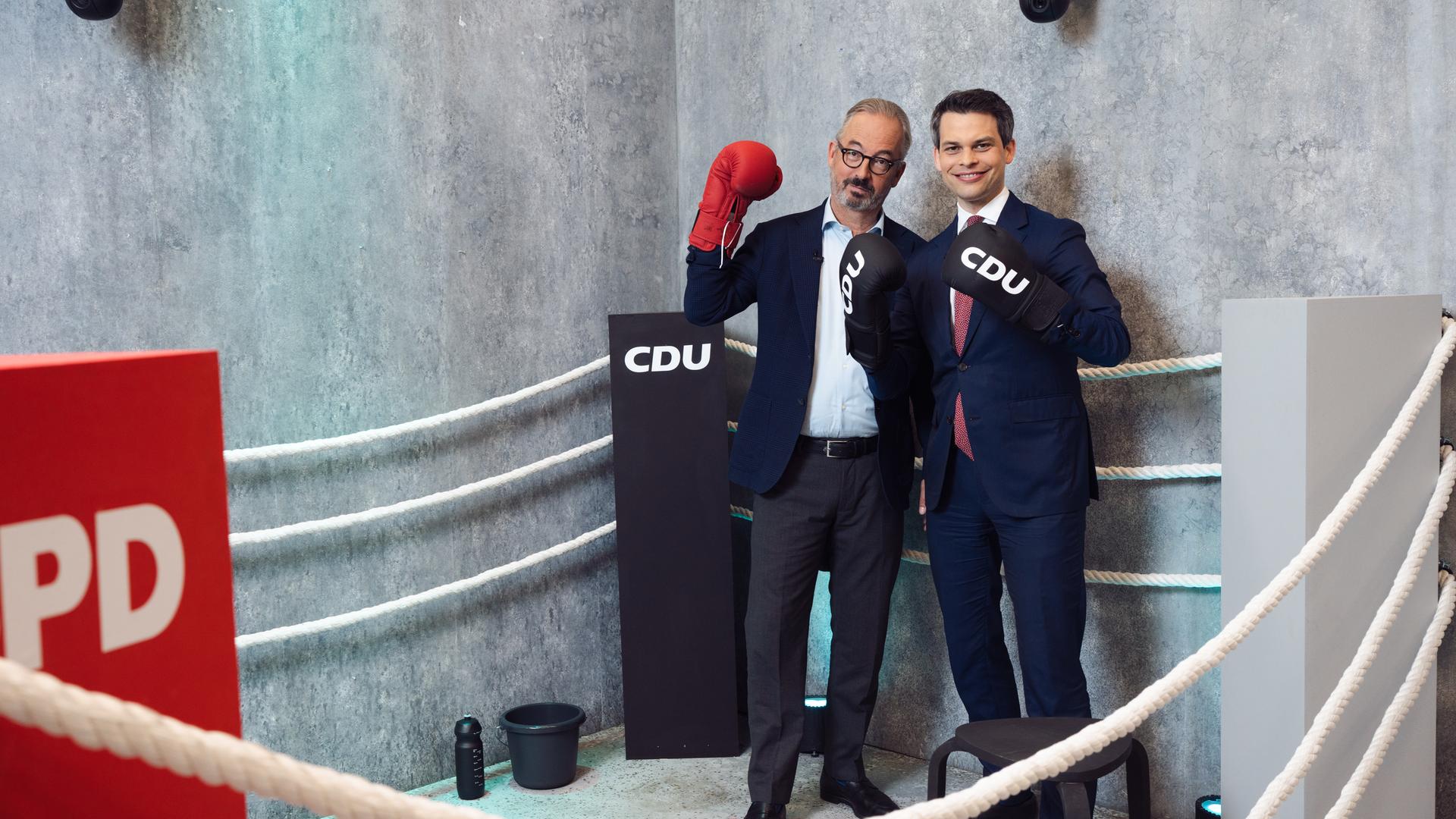 Das Bild zeigt Jan Fleischhauer und den CDU-Politiker Christoph Ploß in einem Boxring-Setting. Beide stehen nebeneinander innerhalb eines Rings mit weißen Seilen vor einer grauen Betonwand. Jan Fleischhauer trägt einen dunkelblauen Anzug mit hellem Hemd und Brille und hält einen roten Boxhandschuh hoch. Christoph Ploß steht rechts neben ihm im dunklen Anzug mit weißem Hemd und roter Krawatte. Er trägt schwarze Boxhandschuhe mit dem Aufdruck „CDU“ und lächelt direkt in die Kamera.
