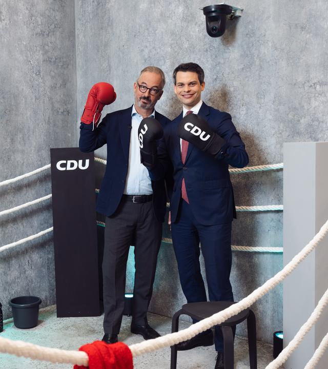 Das Bild zeigt Jan Fleischhauer und den CDU-Politiker Christoph Ploß in einem Boxring-Setting. Beide stehen nebeneinander innerhalb eines Rings mit weißen Seilen vor einer grauen Betonwand. Jan Fleischhauer trägt einen dunkelblauen Anzug mit hellem Hemd und Brille und hält einen roten Boxhandschuh hoch. Christoph Ploß steht rechts neben ihm im dunklen Anzug mit weißem Hemd und roter Krawatte. Er trägt schwarze Boxhandschuhe mit dem Aufdruck „CDU“ und lächelt direkt in die Kamera.