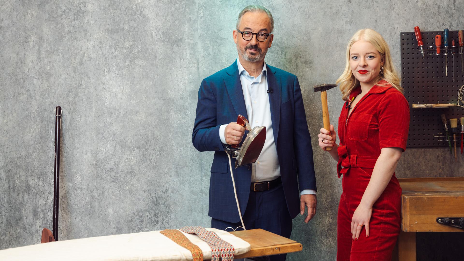 "Keine Talkshow - Eingesperrt mit Jan Fleischhauer": Inszenierte Szene in einem Raum mit grauer Betonwand und industriellem Charakter: Jan Fleischhauer hält ein Bügeleisen über einem Bügelbrett, auf dem mehrere Krawatten liegen. Neben ihm steht Jette Nietzard und hält einen Hammer in der Hand. Beide schauen in die Kamera. Hinter ihnen steht eine Werkbank, über der eine Lochwand mit Werkzeugen hängt.