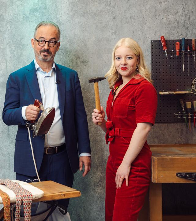 "Keine Talkshow - Eingesperrt mit Jan Fleischhauer": Inszenierte Szene in einem Raum mit grauer Betonwand und industriellem Charakter: Jan Fleischhauer hält ein Bügeleisen über einem Bügelbrett, auf dem mehrere Krawatten liegen. Neben ihm steht Jette Nietzard und hält einen Hammer in der Hand. Beide schauen in die Kamera. Hinter ihnen steht eine Werkbank, über der eine Lochwand mit Werkzeugen hängt.
