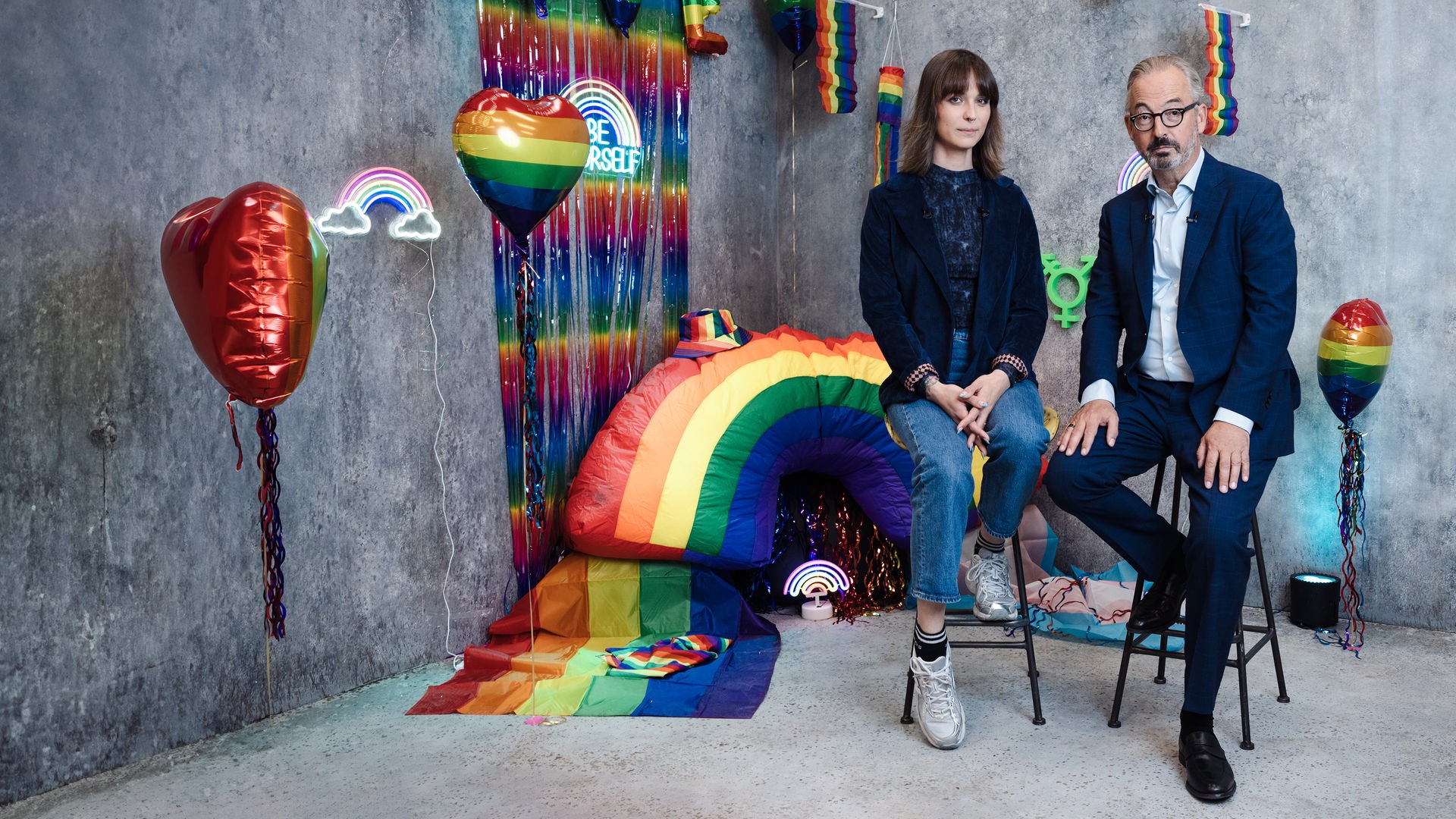 Jan Fleischhauer und Nyke Sławik sitzen auf hohen Hockern vor einer grauen Studiokulisse, die mit Regenbogenfarben und verschiedenen dekorativen Elementen gestaltet ist. Zu sehen sind unter anderem Ballons, kleine Regenbogenflaggen, eine leuchtende Dekoration sowie ein großes Regenbogenobjekt im Hintergrund. Beide blicken in die Kamera. Jan Fleischhauer trägt einen dunklen Anzug mit hellem Hemd, Nyke Sławik ein dunkles Oberteil mit Jeans und Sneakers. Die Szene wirkt wie ein thematisch inszeniertes Studio-Setting.