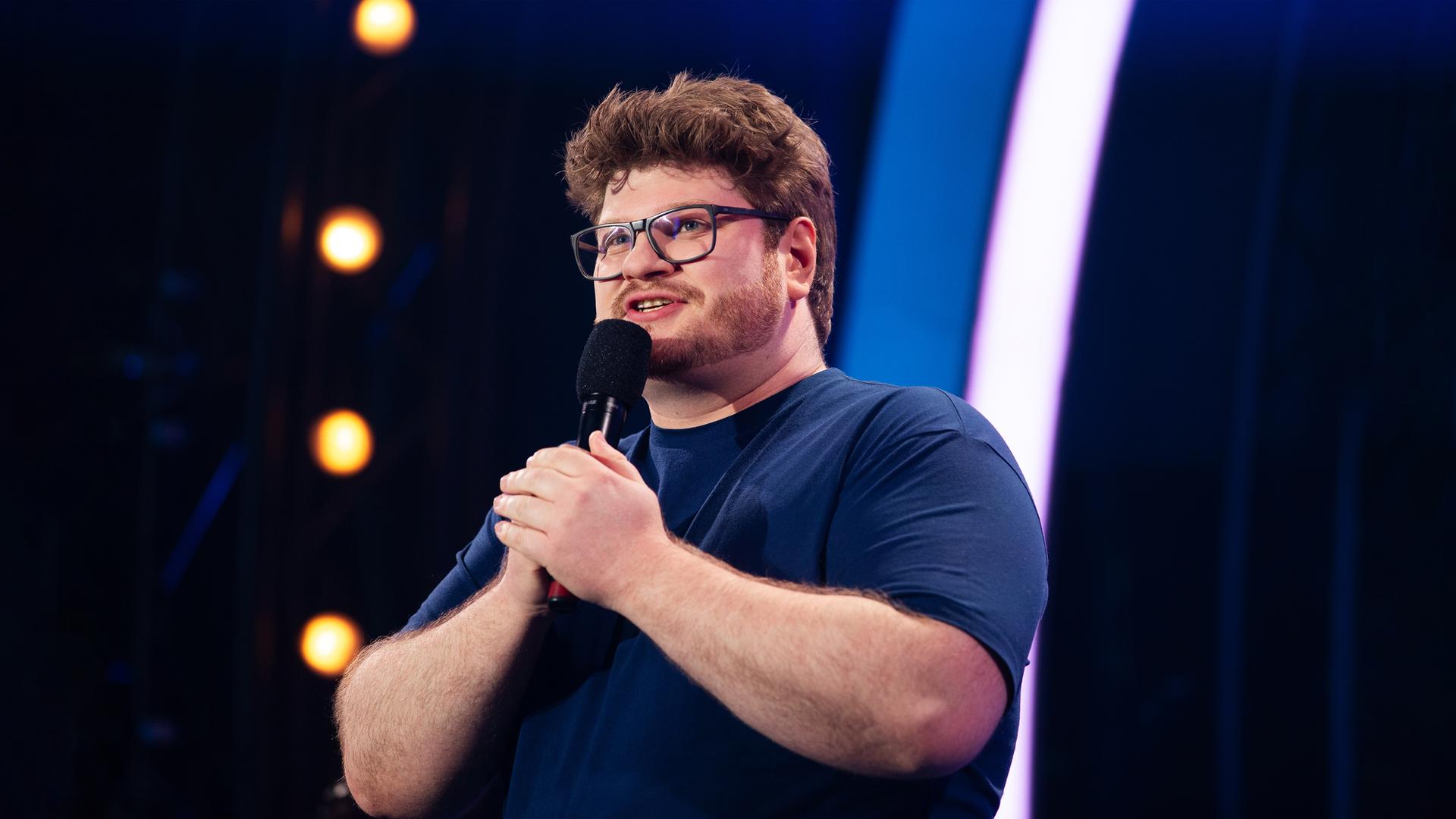 Jan Philipp Zymny steht in der Mitte des Bildes. Er hält mit beiden Händen das Mikrofon fest. Der Hintergrund ist blau beleuchtet und er trägt ein dunkelblaues T-Shirt.