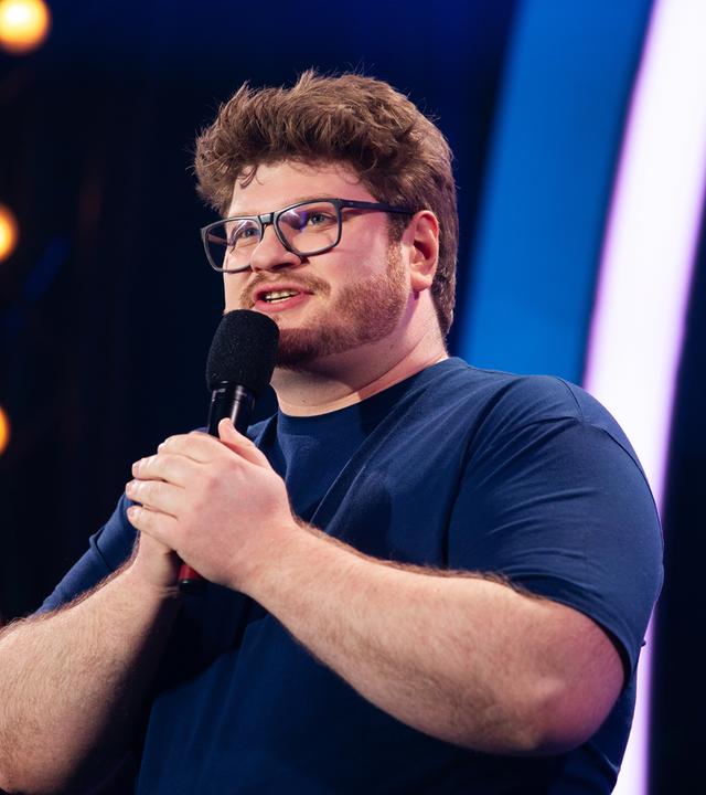 Jan Philipp Zymny steht in der Mitte des Bildes. Er hält mit beiden Händen das Mikrofon fest. Der Hintergrund ist blau beleuchtet und er trägt ein dunkelblaues T-Shirt.