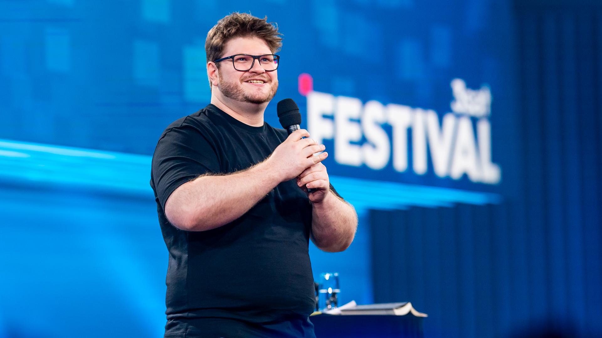 Jan Philipp Zymny steht in der Mitte des Bildes. Er lacht und hält sein Mikrofon mit beiden Händen fest. Das Licht auf der Bühne ist blau. Er trägt ein schwarzes Oberteil.