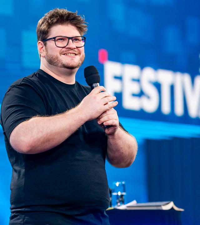 Jan Philipp Zymny steht in der Mitte des Bildes. Er lacht und hält sein Mikrofon mit beiden Händen fest. Das Licht auf der Bühne ist blau. Er trägt ein schwarzes Oberteil.
