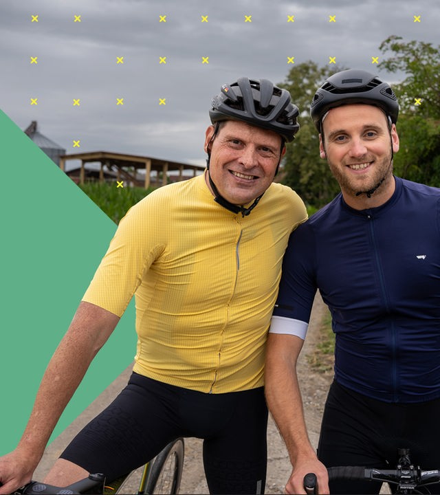 Dr. Leon Windscheid steht lächelnd neben Jan Ullrich auf einem Feldweg. Beide tragen Sportkleidung und Fahrradhelme. Im Hintergrund ist ein Maisfeld und ein grauer Himmel zu sehen.
