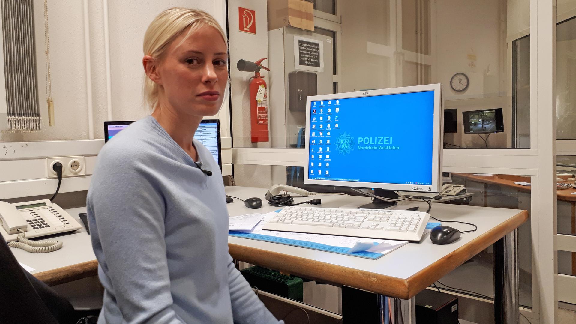 Jana L. sitzt auf einem Schreibtischstuhl, der Kamera zugewandt, hinter ihr ein Computerbildschirm mit einem Desktop-Hintergrund, auf dem "Polizei Nordrhein-Westfalen" steht.