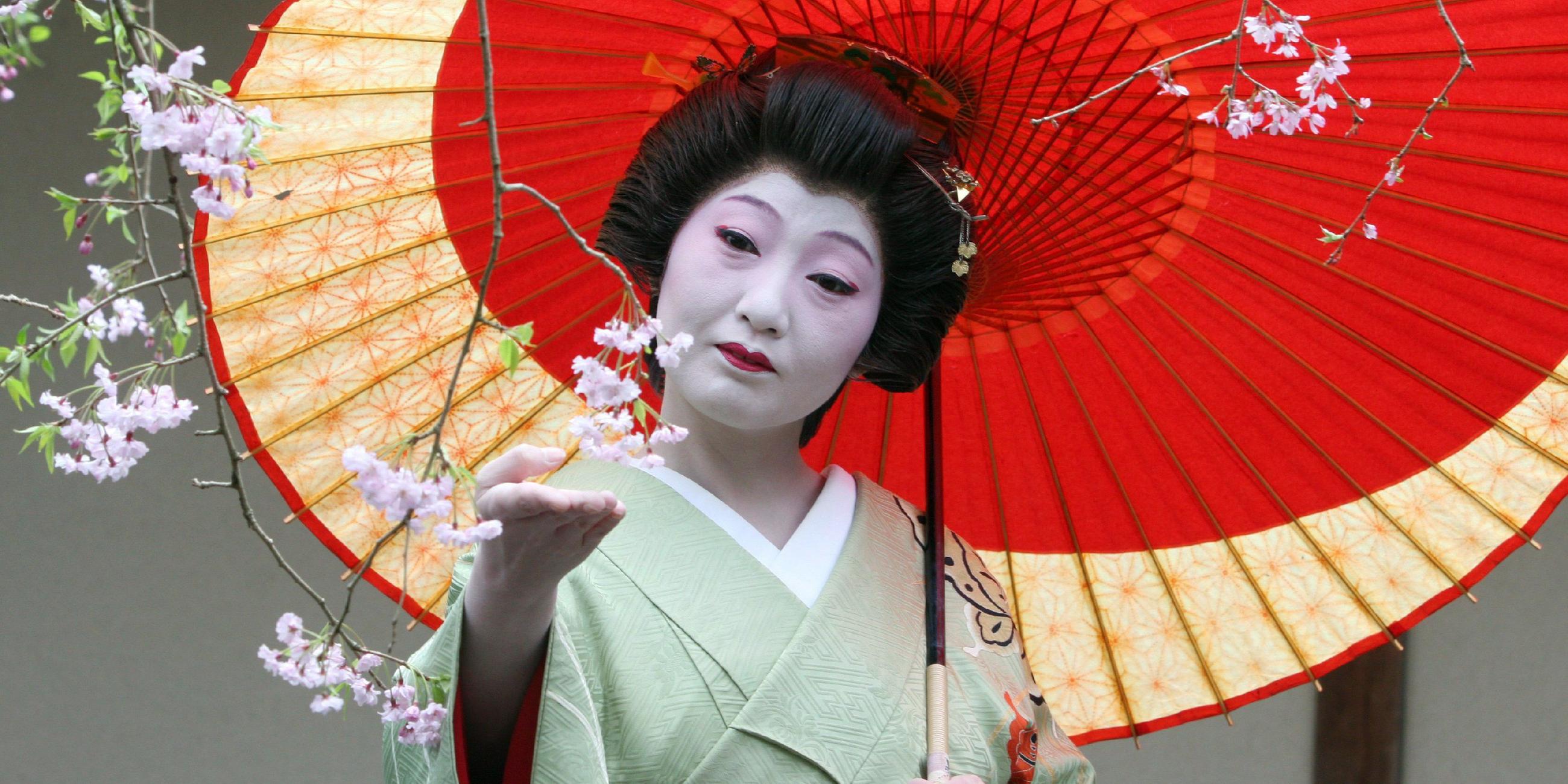 Eine japanische Geisha betrachtet den Ast eines japanischen Kirschbaums. Sie hält einen dekorativen, roten Regenschirm.