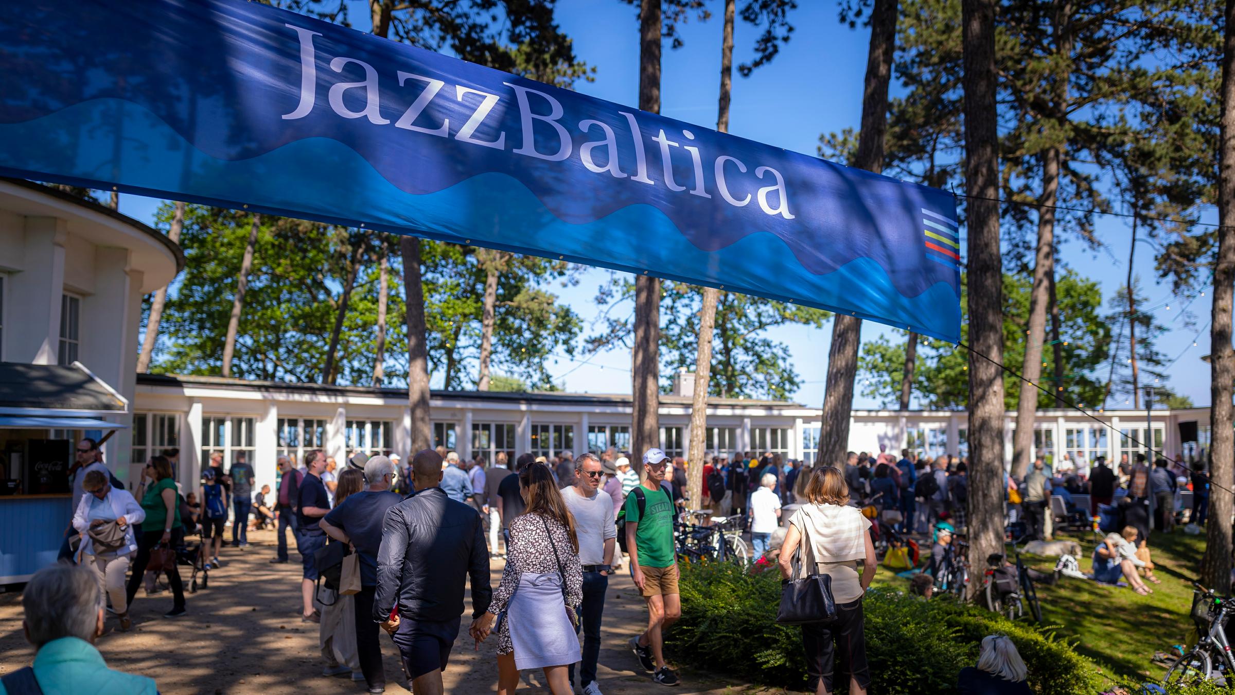 Eingangsbereich zur "Jazz Baltica 2019". Banner der Veranstaltung zwischen zwei hohen Kiefern; darunter Besucher; im Hintergrund ein flaxcxhes Gebäude