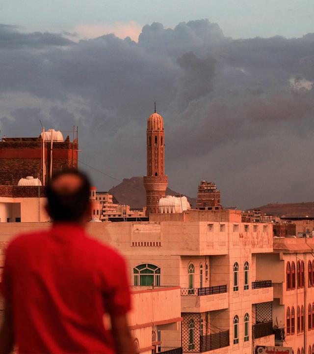 Ein Mann blickt auf ein Minarett, das sich über der Skyline eines Stadtviertels erhebt; aufgenommen bei Sonnenuntergang in Sanaa, der von den Houthis kontrollierten Hauptstadt des Jemen