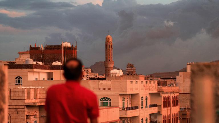 Ein Mann blickt auf ein Minarett, das sich über der Skyline eines Stadtviertels erhebt; aufgenommen bei Sonnenuntergang in Sanaa, der von den Houthis kontrollierten Hauptstadt des Jemen