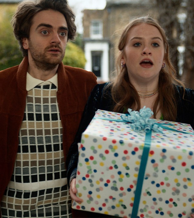 "Extraordinary - Jen vs. Nora: Der ultimative Showdown": Jizzlord (Luke Rollason) und Jen (Máiréad Tyers) stehen vor der Türe. Im Hintergrund stehen Häuser einer schicken Wohngegend. Jen hält ein großes Geschenk, das mit gepunktetem Papier und blauer Schleife eingewickelt ist, in den Händen. Jizzlord schaut erstaunt.