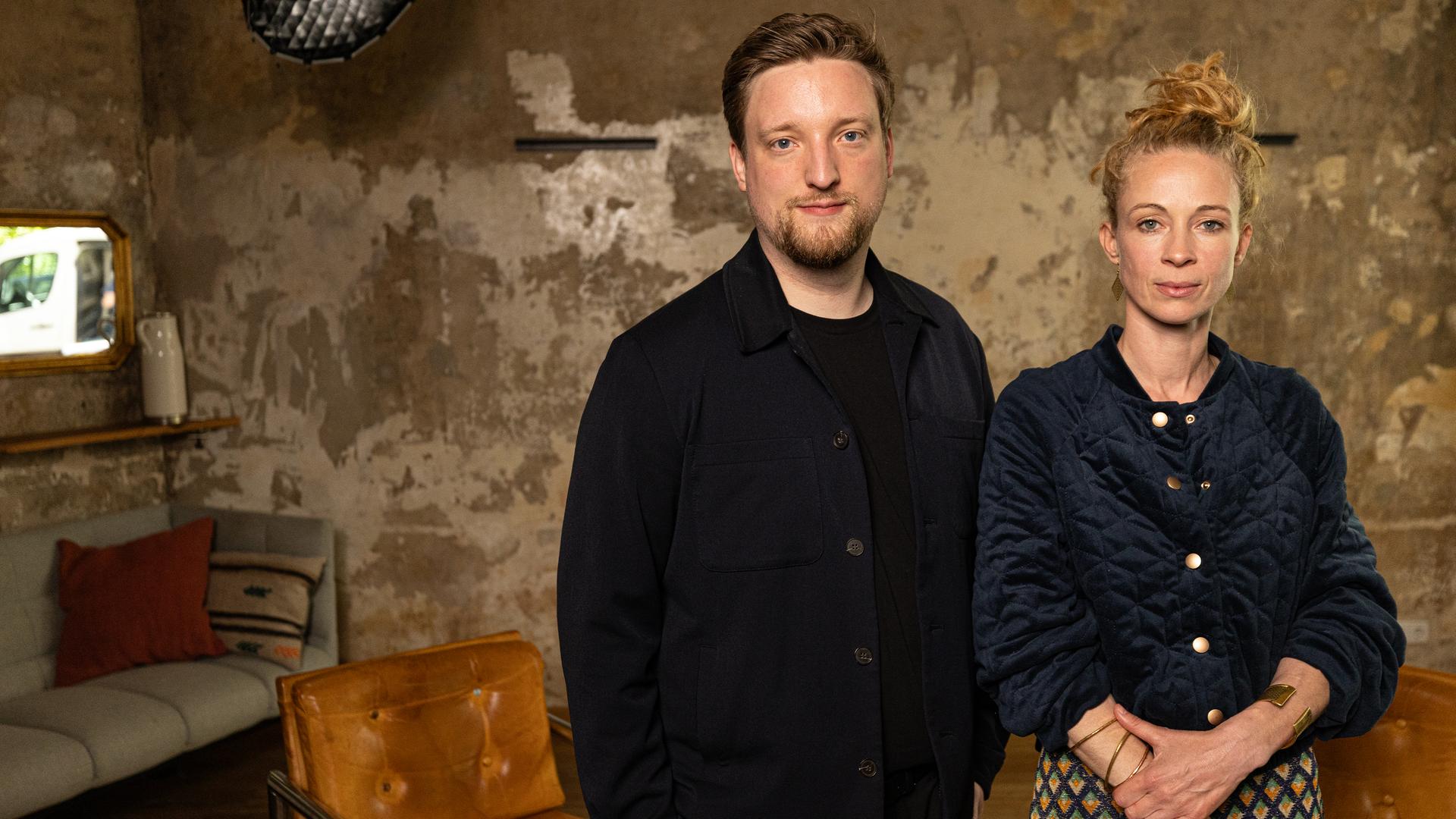 Ein Mann (Jens Teutrine) und eine Frau (Helena Steinhaus) stehen nebeneinander vor einer Wand an der die Tapete abblättert und fehlt. Im Hintergrund ist auch noch ein Sofa zu sehen.
