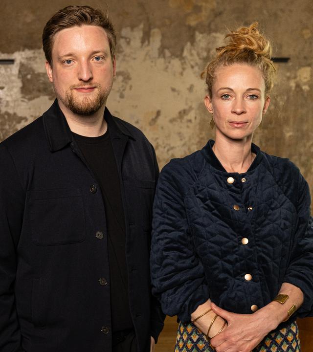 Ein Mann (Jens Teutrine) und eine Frau (Helena Steinhaus) stehen nebeneinander vor einer Wand an der die Tapete abblättert und fehlt. Im Hintergrund ist auch noch ein Sofa zu sehen.