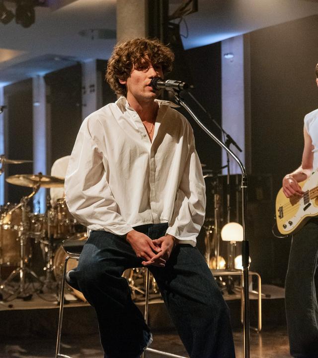 Der Sänger von Jeremias sitzt auf einem Hocker und singt ins Mikrofon. Im Hintergrund ist ein Schlagzeug zu sehen und ein weiterer junger Mann, der Bass spielt.