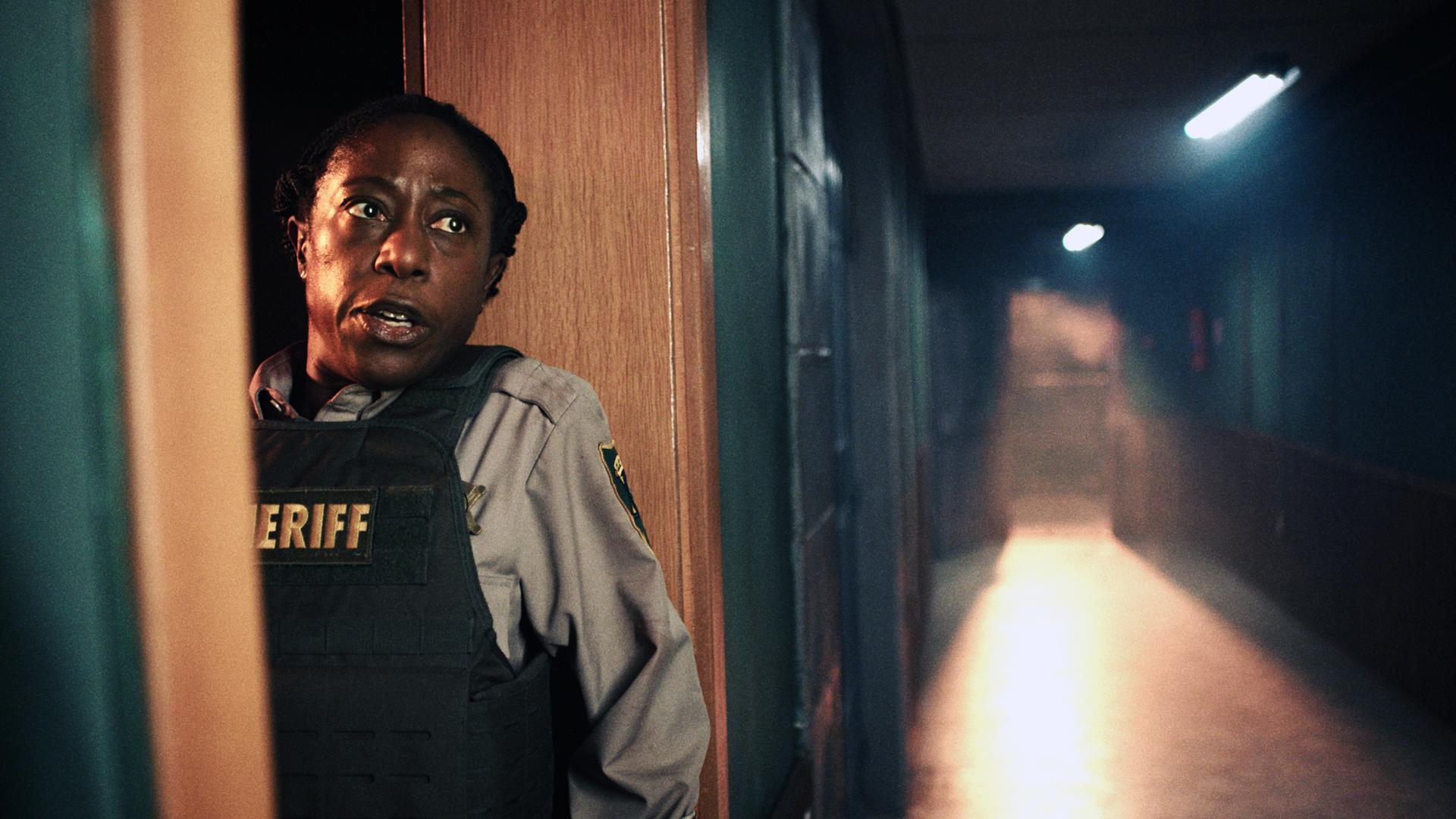 Tabby (Nikki Amuka-Bird) ist angsterfüllt und versteckt sich hinter einem Türrahmen. Der Gang neben ihr ist leer.