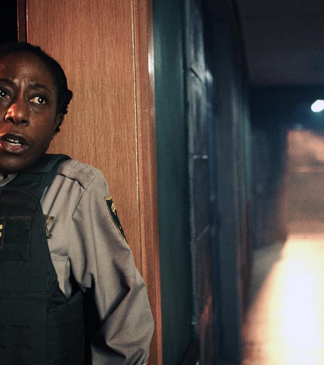 Tabby (Nikki Amuka-Bird) ist angsterfüllt und versteckt sich hinter einem Türrahmen. Der Gang neben ihr ist leer.