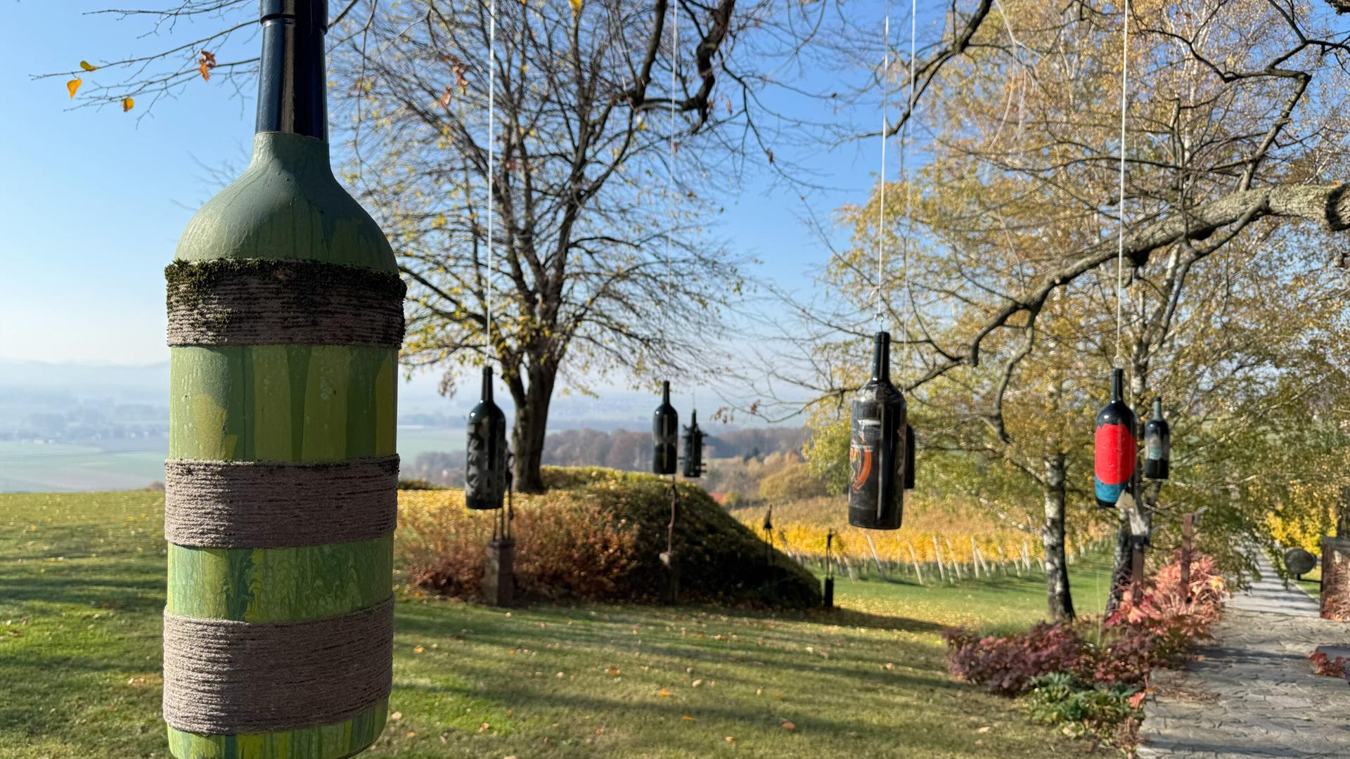 Das Bild zeigt eine ländliche Weinanbauumgebung in Slowenien, wahrscheinlich in der Region Jeruzalem. Im Vordergrund hängt eine große grüne Weinflasche, die teilweise bemalt und mit einem Faden umwickelt ist. Im Hintergrund und an den Ästen eines Baumes hängen mehrere weitere Weinflaschen in verschiedenen Farben, darunter schwarz, rot und blau. Die Flaschen sind gleichmäßig zwischen den Zweigen angeordnet und schwingen sanft im Wind.  Die Umgebung ist von sanften Hügeln und Weinreben umgeben. Der Himmel ist klar und blau, mit wenigen Wolken. In der Ferne sind weitere Weinreben sichtbar, die sich über die Hügel erstrecken. Der Boden ist grün und gepflegt, und der Weg ist gepflastert, der eine ruhige, ländliche Atmosphäre schafft.  Insgesamt vermittelt das Bild eine harmonische Verbindung zwischen Natur und Weinbau, die typisch für diese Region ist.