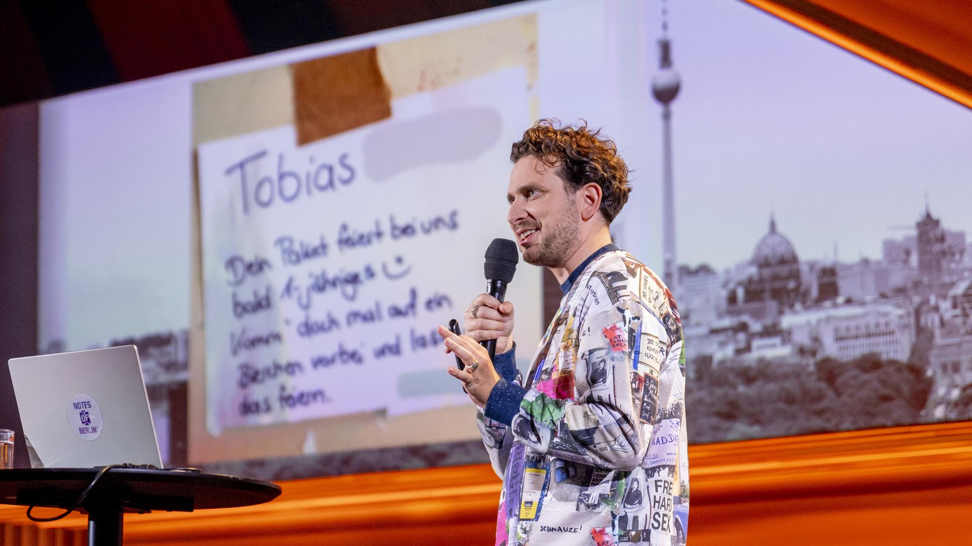 Joab Nist steht auf der rechten Seite des Bildes. Auf der linken Bildseite steht ein Laptop auf einem Stehtisch. Im Hintergrund ist ein Bild der Berliner Skyline zu sehen. Er trägt einen bunten Pullover und hält das Mikrofon in seiner rechten Hand.