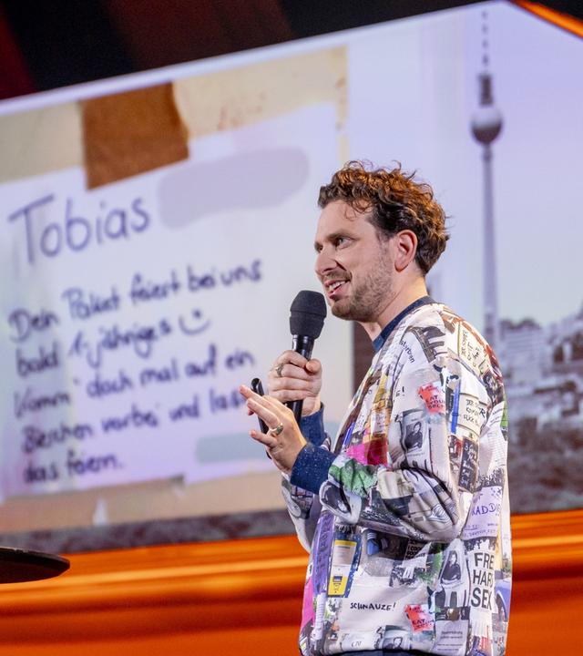 Joab Nist steht auf der rechten Seite des Bildes. Auf der linken Bildseite steht ein Laptop auf einem Stehtisch. Im Hintergrund ist ein Bild der Berliner Skyline zu sehen. Er trägt einen bunten Pullover und hält das Mikrofon in seiner rechten Hand.
