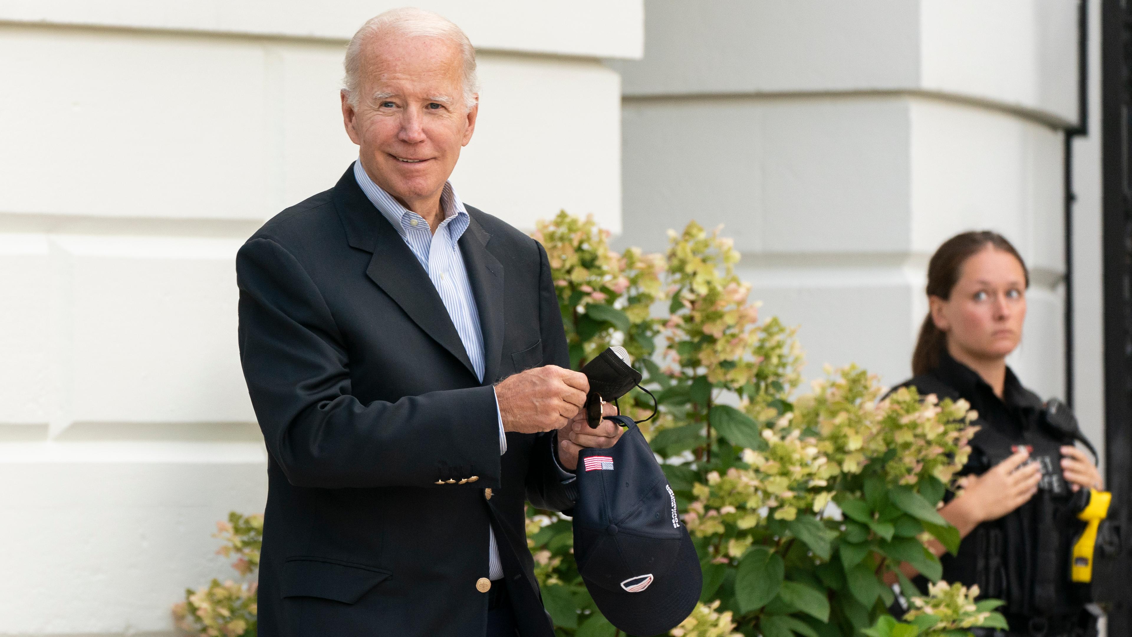 Joe Biden tritt aus einer Tür und zieht sich grinsend seinen Mund-Nasen-Schutz vom Gesicht (Archivbild).