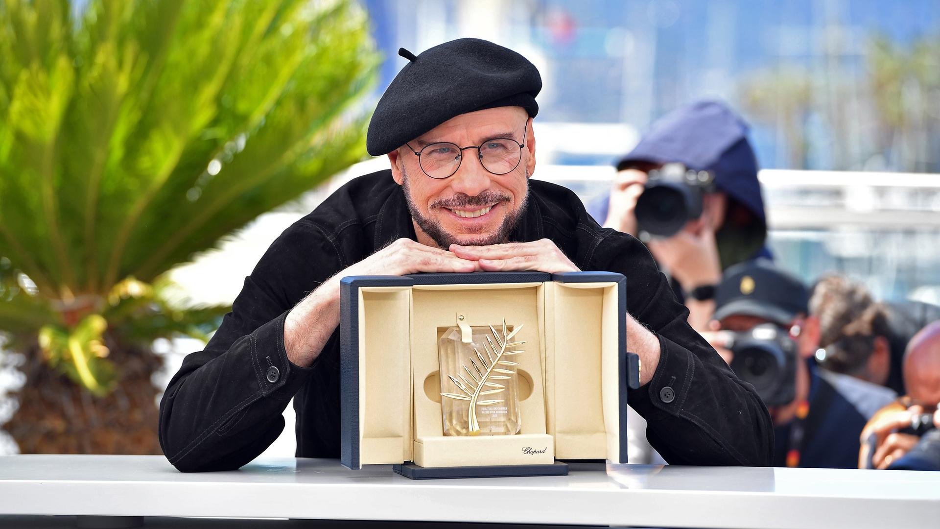 Der Schauspieler John Travolta, ein schwarz gekleideter Mann mit Baskenmütze, brille und Bart stützt sich lächelnd auf der goldenen Ehrenpalme von Cannes ab, einer goldenen Trophäe in einer beige ausgeschlagenen Schatulle.