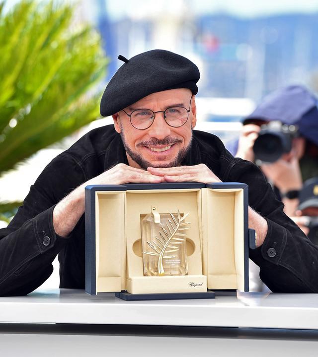 Der Schauspieler John Travolta, ein schwarz gekleideter Mann mit Baskenmütze, brille und Bart stützt sich lächelnd auf der goldenen Ehrenpalme von Cannes ab, einer goldenen Trophäe in einer beige ausgeschlagenen Schatulle.