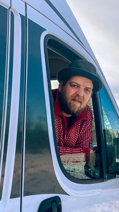 "Jonny Island": Ein Mann mit Hut und Bart guckt aus dem Fenster eines Wohnmobils. Im Hintergrund eine leere winterliche Landstraße.