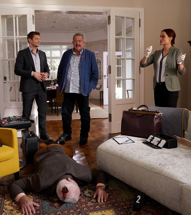 "Die Rosenheim-Cops - Der letzte Jour fixe": Die Kommissare Hansen (Igor Jeftic) und Hofer (Joseph Hannesschläger) stehen mit Pathologin Mai (Sina Wilke) im Wohnzimmer eines Hauses. Auf dem Teppich vor ihnen, zwischen Sessel und Couch, liegt das Mordopfer auf dem Bauch; am Hinterkopf ist eine blutende Wunde zu sehen. Ein Kollege der KTU (Komparse) sichert Spuren an einer Tür.