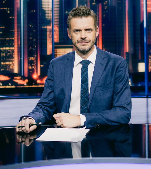 Florian Schroeder sitzt am Schreibtisch im Studio seiner neuen Show "JOURNAL DELUXE". Im Hintergrund ist durch ein ovales Fenster eine Skyline mit Hochhäusern zu sehen.