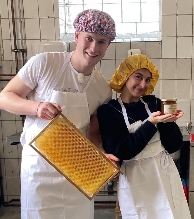 Die WG - Das große Wiedersehen: Mark und Ella aus der Jubiläums-WG stehen in einer weiß gekachelten Industrieküche. Beide tragen aus hygienischen Gründen weiße Schürzen und eine Haube über den Haaren. Ella präsentiert in ihren Händen ein Glas Honig. Mark hält einen Holzrahmen mit Bienenwaben in die Kamera. Beide lächeln.