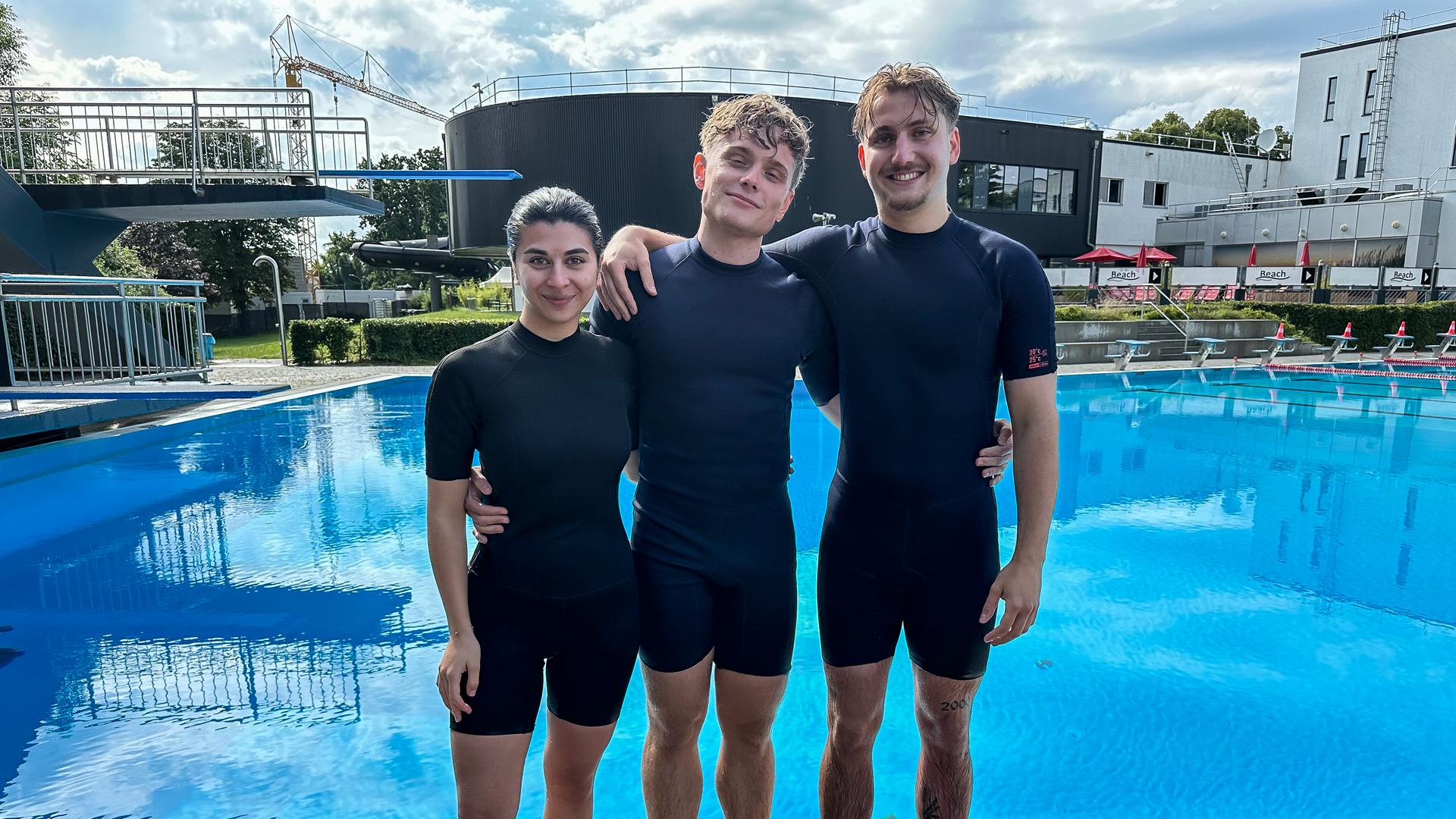 Die WG - Das große Wiedersehen: Ella, Julius und Robin aus der Jubiläums-WG stehen vor einem Schwimmbecken. Alle drei tragen kurze Neoprenanzüge und haben nasse Haare. Sie lächeln in die Kamera und haben sich gegenseitig die Arme um Schultern und Hüften gelegt. Links neben der Gruppe sieht man zwei Sprungtürme.