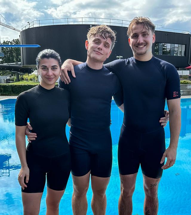 Die WG - Das große Wiedersehen: Ella, Julius und Robin aus der Jubiläums-WG stehen vor einem Schwimmbecken. Alle drei tragen kurze Neoprenanzüge und haben nasse Haare. Sie lächeln in die Kamera und haben sich gegenseitig die Arme um Schultern und Hüften gelegt. Links neben der Gruppe sieht man zwei Sprungtürme.