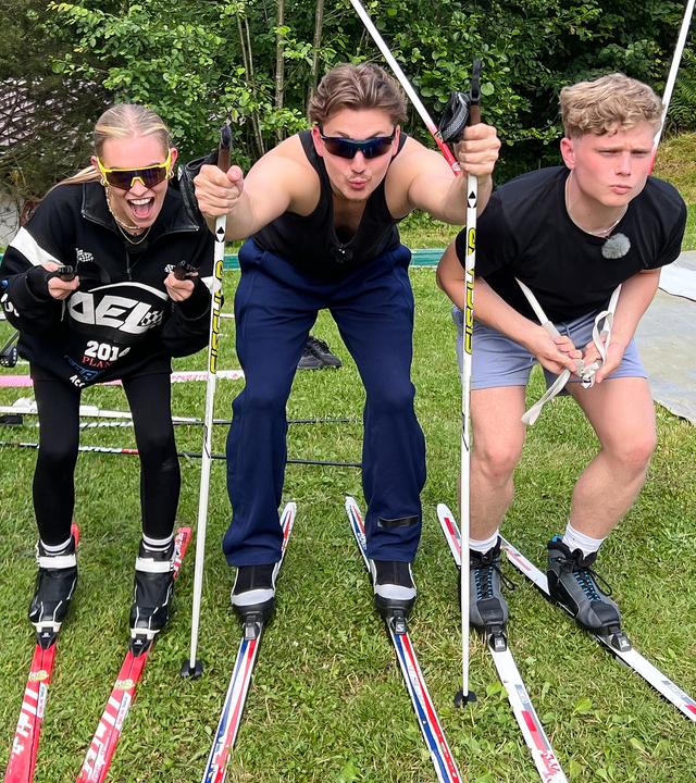Die WG - Das große Wiedersehen: lexa, Robin und Julius stehen nebeneinander auf einer grünen Wiese und tragen Langlaufskier und Stöcke. Sie tun so, als würden sie mit Skiern gleich losfahren wollen. Alexa und Julius haben sich die Stöcke dafür unter die Arme geklemmt, Robin hat beide Stöcke in den Boden gesteckt, um sich vermeintlich abzustoßen. Alexa und Robin tragen Sonnenbrillen und lange Trainingshosen, Julius steht scheinbar konzentriert mit kurzer Hose und T-Shirt in Startposition.