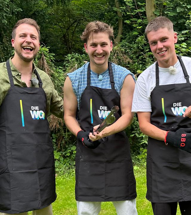 Die WG - Das große Wiedersehen: Jonas, Robin und Jeppe aus der Jubiläums-WG stehen in einem Garten mit Bäumen auf einer Wiese. Sie tragen alle schwarze Grillschürzen mit dem WG-Logo darauf. Robin, in der Mitte der Gruppe, hält eine Grillzange in den Händen. Jeppe, auf der rechten Seite im Bild, zieht Grillhandschuhe an. Alle lachen in die Kamera.