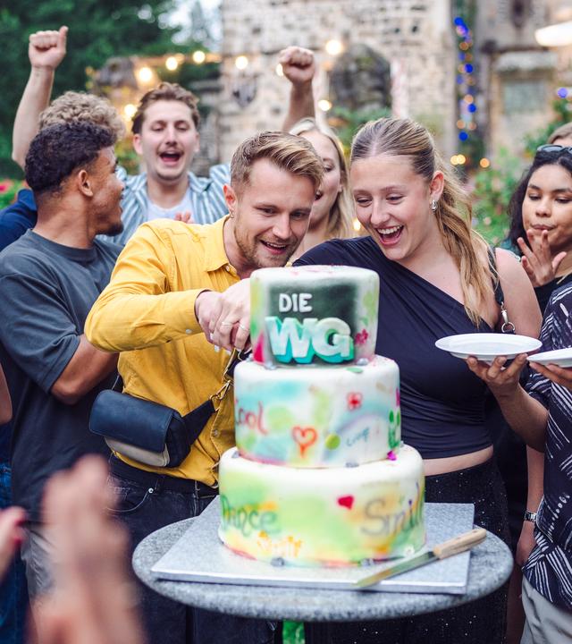 Die WG - Das große Wiedersehen: Im Mittelpunkt des Fotos steht eine dreistöckige, bunte Torte mit WG Logo. Aaron und Hanna aus der Jubiläums-WG stehen hinter der Torte und schneiden sie gerade an. Dominik steht rechts neben Hanna und hält erwartungsvoll zwei Kuchenteller bereit. Viele andere WGlerinnen und WGler stehen um die Gruppe herum, klatschen und jubeln und freuen sich offenbar auf die Torte. Im Hintergrund ist die Burg zu sehen, in der die Jubiläums-WG stattgefunden hat.
