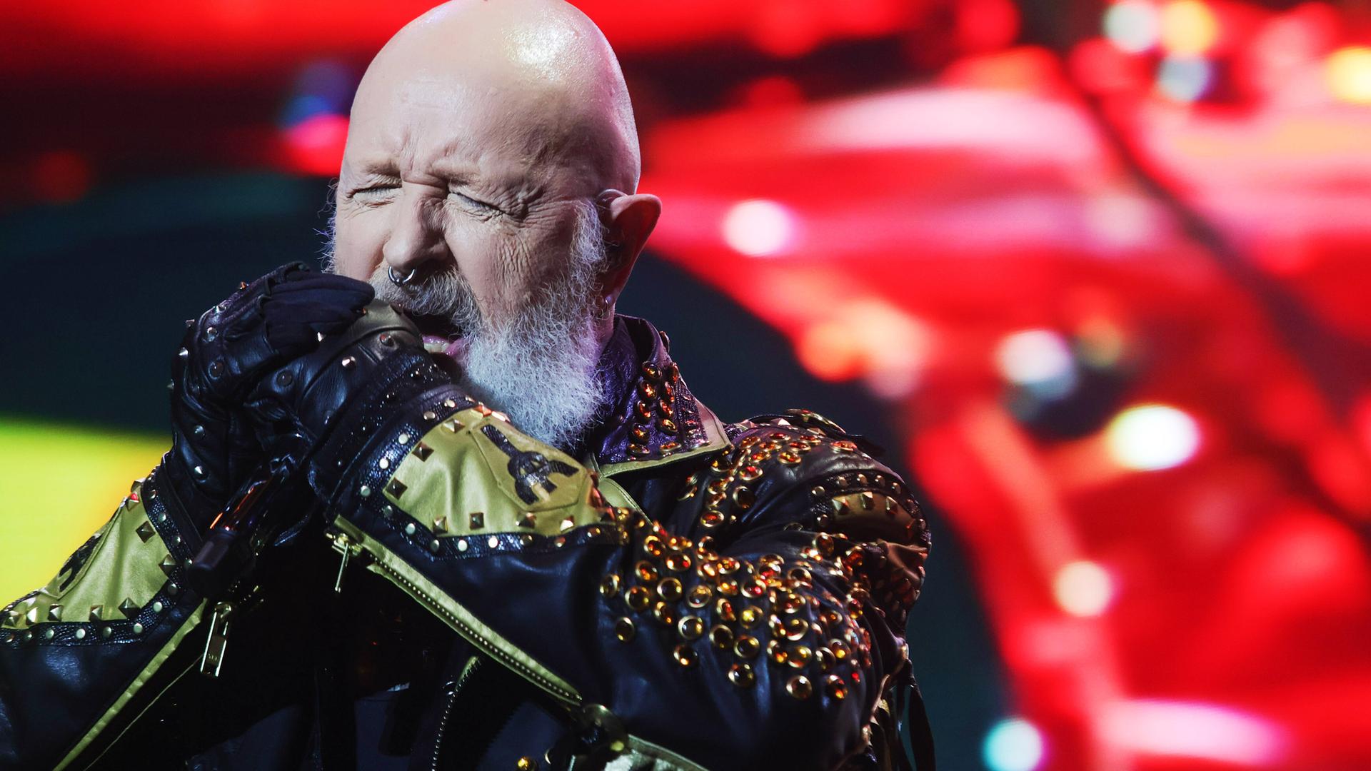 Ein älterer Mann mit Glatze und weißem Bart trägt ein Lederkostüm mit vielen Nieten und singt auf der Bühne in ein Mikrofon.