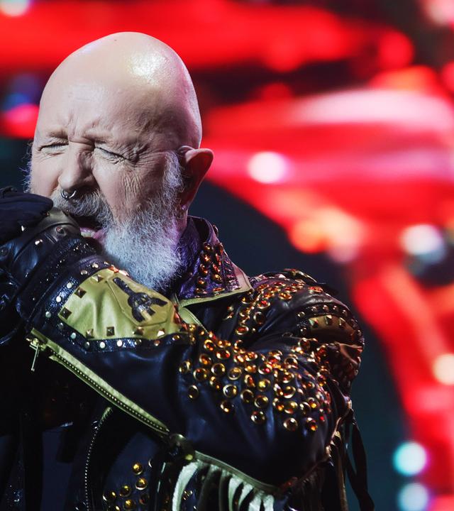 Ein älterer Mann mit Glatze und weißem Bart trägt ein Lederkostüm mit vielen Nieten und singt auf der Bühne in ein Mikrofon.