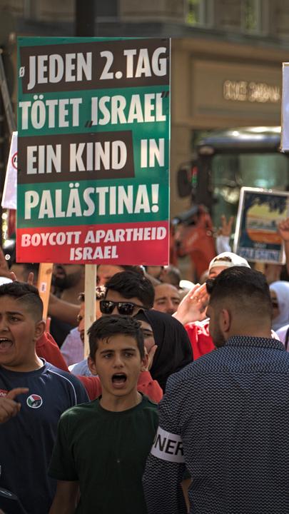 Während einer Demonstration halten mehrere Menschen Schilder in die Luft und schreien. Auf einem der Schilder steht die Aufschrift "Jeden 2. Tag tötet Israel ein Kind in Palästina".