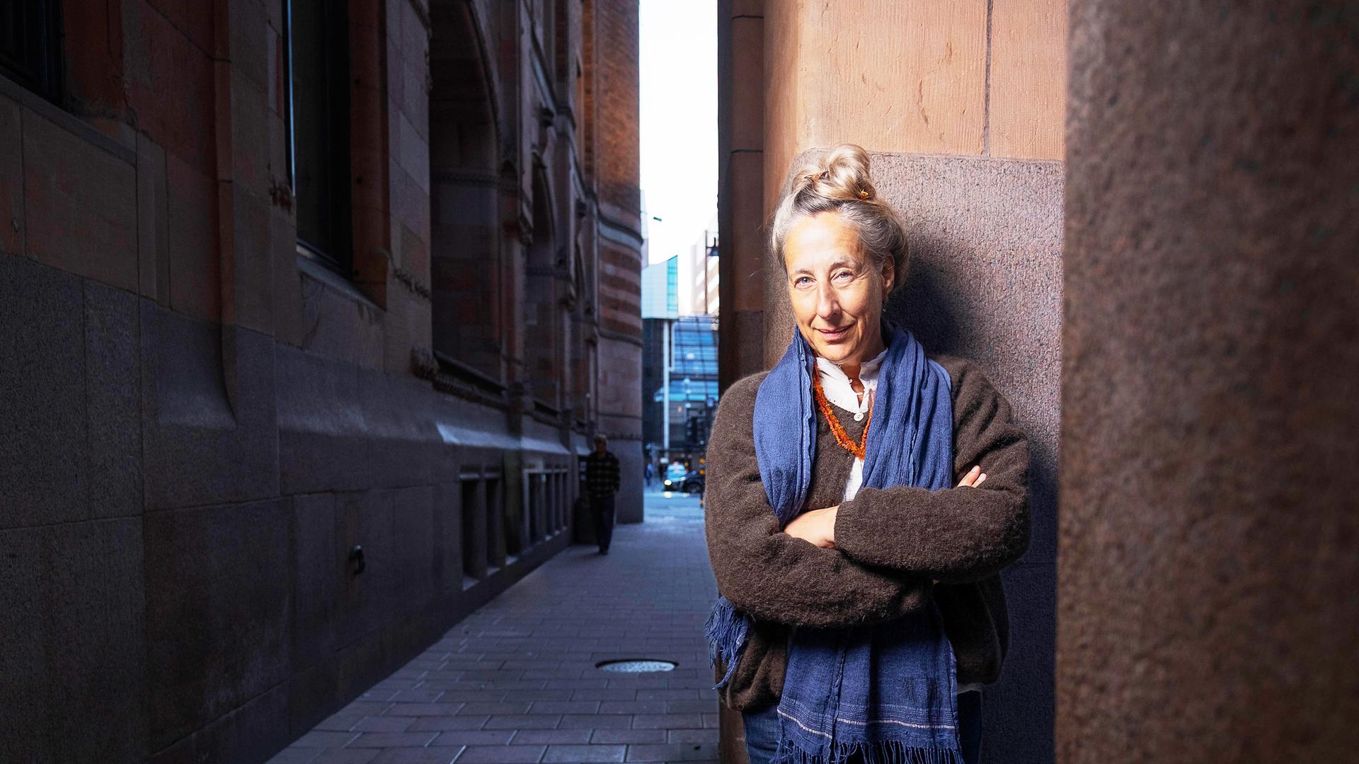 Eine Frau mittleren Alters mit hochgesteckten graublonden Haaren und bekleidet mit einem Strickpullover und Schal steht an einer Straßenecke und lächelt in die Kamera.