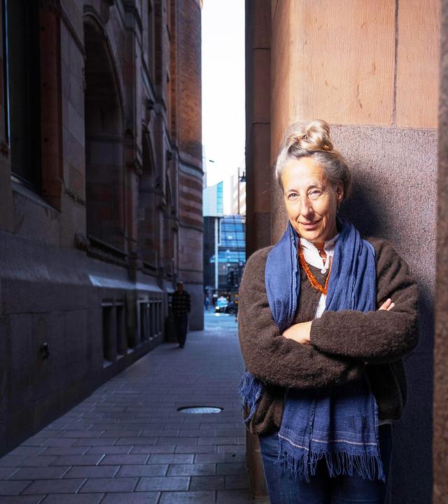 Eine Frau mittleren Alters mit hochgesteckten graublonden Haaren und bekleidet mit einem Strickpullover und Schal steht an einer Straßenecke und lächelt in die Kamera.