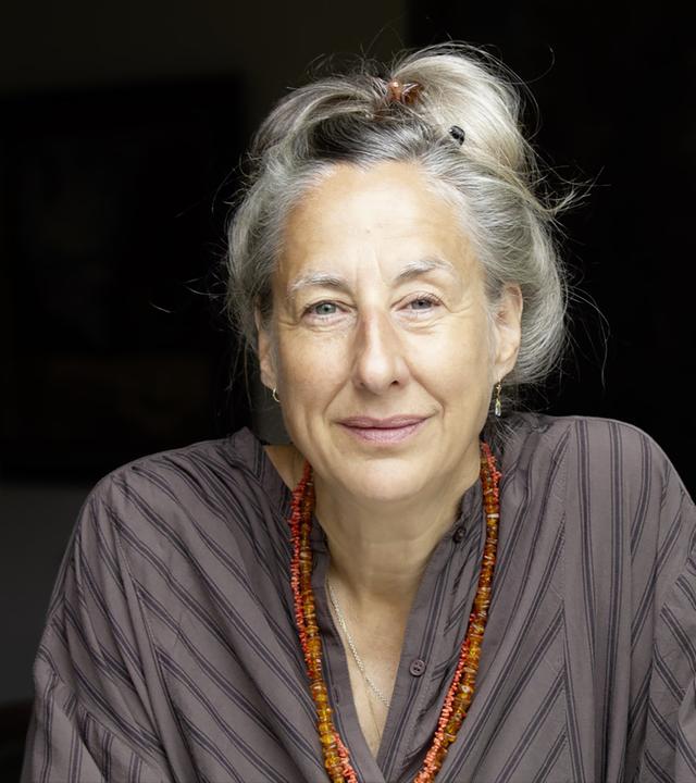 Porträtfoto einer Frau mittleren Alters mit hochgesteckten graublonden Haaren, die eine lange doppelreihige orangefarbene Edelsteinkette trägt.